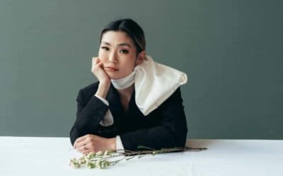 Asian woman with scarf wrapped around her neck sitting, elbow on table with her hand resting on her cheek.
