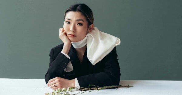 Asian woman with scarf wrapped around her neck sitting, elbow on table with her hand resting on her cheek.