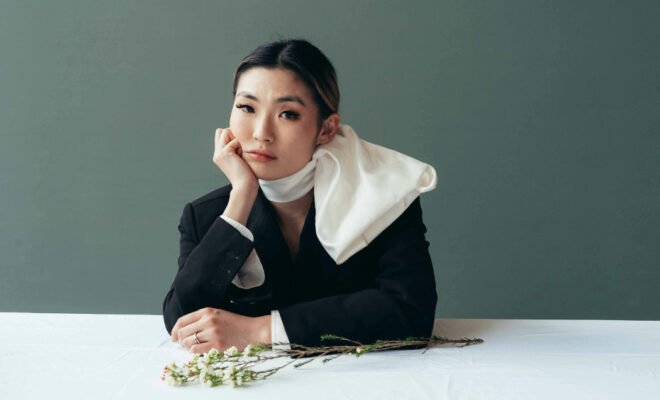 Asian woman with scarf wrapped around her neck sitting, elbow on table with her hand resting on her cheek.