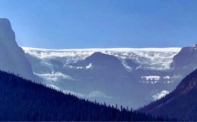 The image is of a long white glacier on top of the Rocky Mountains, high above the alpine trees.