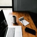 A black wall with a black outlined window open to the outside. A wooden desk table next to the wall housing a laptop, eye glasses and a cup of coffee.