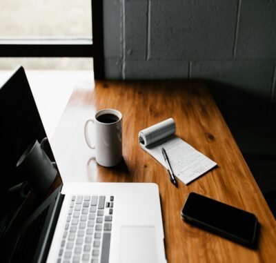 A black wall with a black outlined window open to the outside. A wooden desk table next to the wall housing a laptop, eye glasses and a cup of coffee.