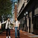 Three friends are walking along side one another near some shops, laughing together as the friend in the middle is reaching out to touch both of the other friends beside them