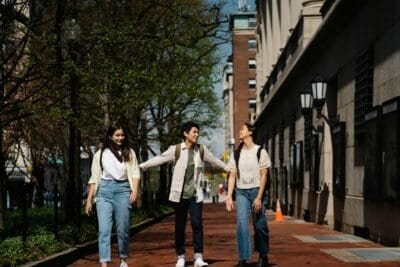 Three friends are walking along side one another near some shops, laughing together as the friend in the middle is reaching out to touch both of the other friends beside them
