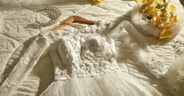 embroidered white lace wedding dress laying on a white bed next to a yellow bouquet