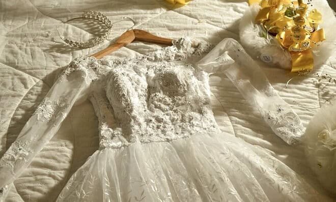 embroidered white lace wedding dress laying on a white bed next to a yellow bouquet
