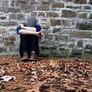 A person sits in front of a brick wall with their knees to their chest, leaves covering the floor around them. They appear sad, deep in their own thoughts