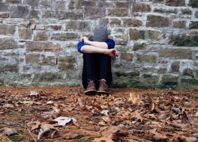 A person sits in front of a brick wall with their knees to their chest, leaves covering the floor around them. They appear sad, deep in their own thoughts