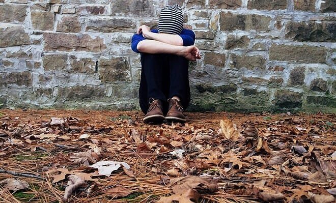 A person sits in front of a brick wall with their knees to their chest, leaves covering the floor around them. They appear sad, deep in their own thoughts