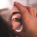 A woman is holding up a small shard from a mirror. In the reflection, it only shows her eye and a slight outline of her nose, as if showing how she ponders with what she notices within the depths of her vision