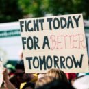 There is a protest going on with people crowded together, though the image is focused on one one specific sign being held up. The sign reads "Fight today for a better tomorrow"