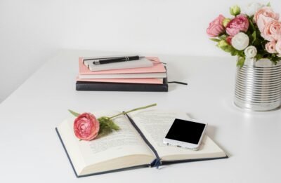 An open book on a white table with an iPhone and pink flower accents