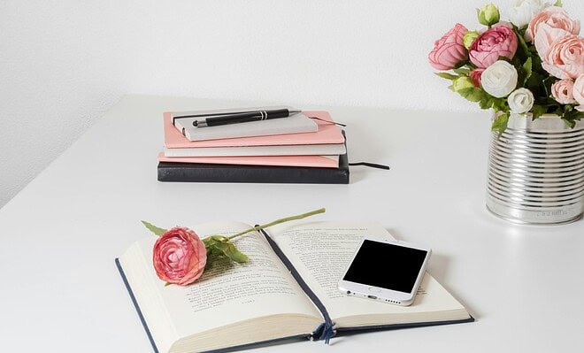 An open book on a white table with an iPhone and pink flower accents