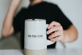 A blurred out woman in the background with a white coffee cup that reads 'see the good.
