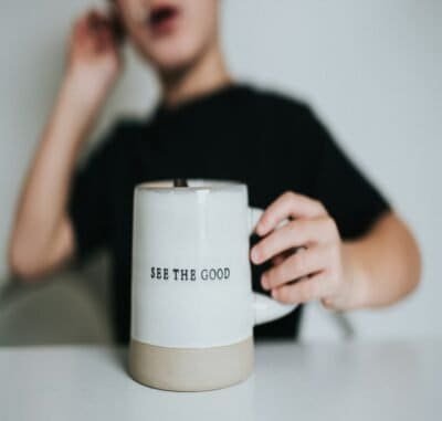 A blurred out woman in the background with a white coffee cup that reads 'see the good.