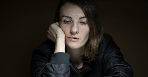 We see a woman sitting alone in a dark room with her hand pressed against her face, showing a depressed expression as she's deep in thought.