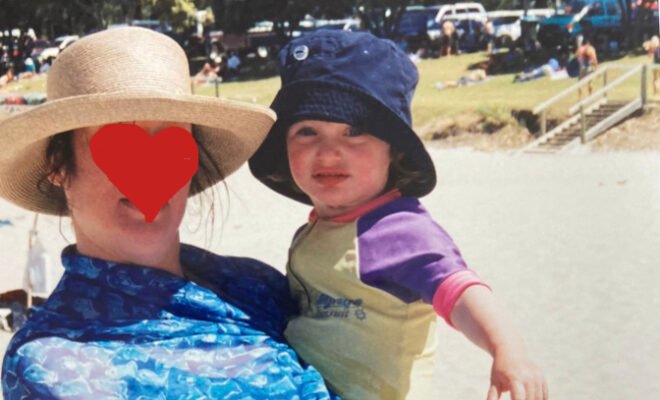 A child stares into camera, held by her mother whose anonymity is preserved through the drawing of a heart over her face.