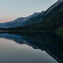 A image of a lakeside view with mountains clear in the background of the lake. The sky is a mix of blue with light shades of orange just peaking out beyond the mountains.