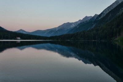 A image of a lakeside view with mountains clear in the background of the lake. The sky is a mix of blue with light shades of orange just peaking out beyond the mountains.