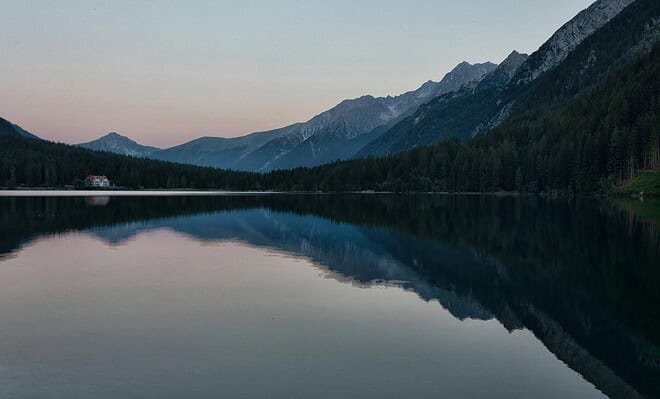 A image of a lakeside view with mountains clear in the background of the lake. The sky is a mix of blue with light shades of orange just peaking out beyond the mountains.