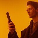 An orange background with a man in a black coat using a flip phone