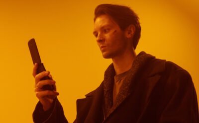 An orange background with a man in a black coat using a flip phone