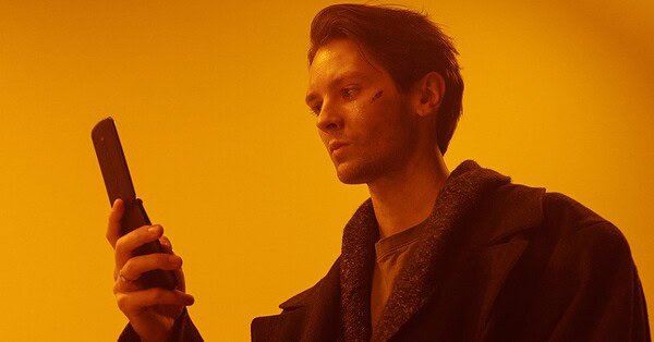 An orange background with a man in a black coat using a flip phone