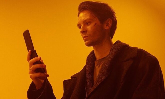 An orange background with a man in a black coat using a flip phone