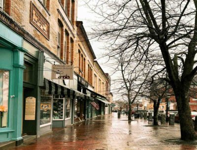 Image of small town Bath, Maine in the rain. Brick shops front a wide sidewalk lined with trees.