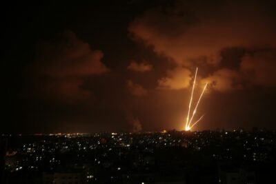 orange and yellow rockets rise into a cloudy night sky over an urban landscape.