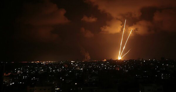 orange and yellow rockets rise into a cloudy night sky over an urban landscape.
