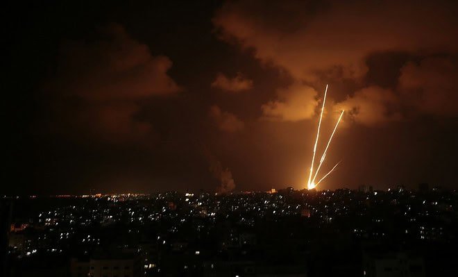 orange and yellow rockets rise into a cloudy night sky over an urban landscape.