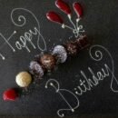White text scrolled on a chalk board that reads "Happy Birthday", interspersed with small shells arranged in a line and as flower buds