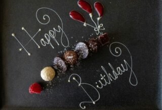 White text scrolled on a chalk board that reads "Happy Birthday", interspersed with small shells arranged in a line and as flower buds