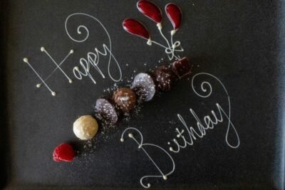 White text scrolled on a chalk board that reads "Happy Birthday", interspersed with small shells arranged in a line and as flower buds