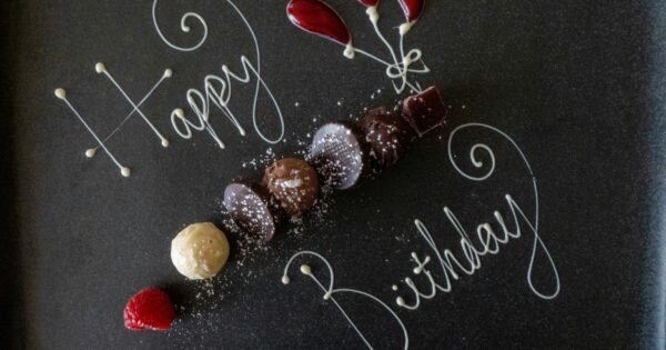 White text scrolled on a chalk board that reads "Happy Birthday", interspersed with small shells arranged in a line and as flower buds