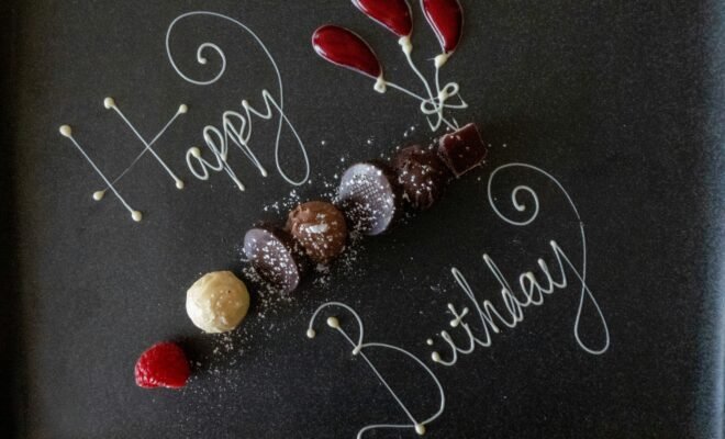 White text scrolled on a chalk board that reads "Happy Birthday", interspersed with small shells arranged in a line and as flower buds