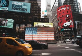There is a view of a busy street in the city, showing different billboards and cars driving past a huge light display of the American flag.
