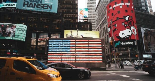 There is a view of a busy street in the city, showing different billboards and cars driving past a huge light display of the American flag.