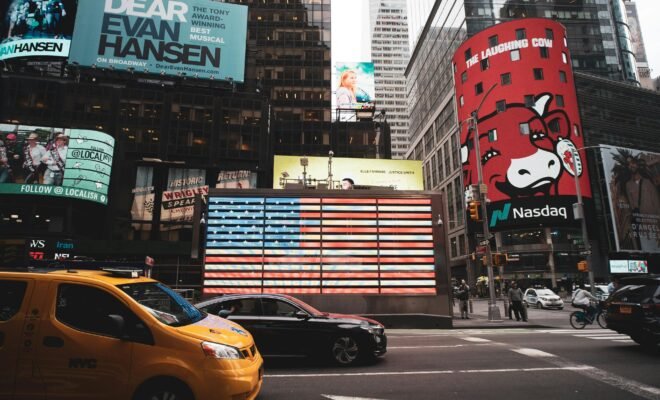 There is a view of a busy street in the city, showing different billboards and cars driving past a huge light display of the American flag.