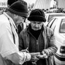 A black and white image of three men in coats and knit caps in front of a line of traffic, in front of the Azadi Tower in Tehran