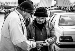 A black and white image of three men in coats and knit caps in front of a line of traffic, in front of the Azadi Tower in Tehran