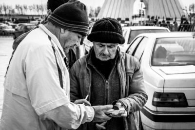 A black and white image of three men in coats and knit caps in front of a line of traffic, in front of the Azadi Tower in Tehran