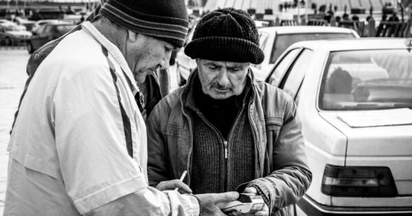 A black and white image of three men in coats and knit caps in front of a line of traffic, in front of the Azadi Tower in Tehran