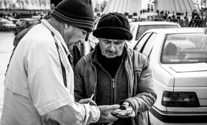A black and white image of three men in coats and knit caps in front of a line of traffic, in front of the Azadi Tower in Tehran