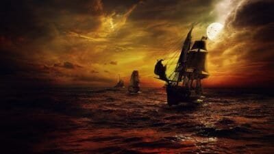 A dark orange and yellow tinted picture fading into black ocean scene with two ships sailing. The one in the foreground is a pirate ship and the smaller one is chasing after the pirate ship. In the background there is a lighthouse.