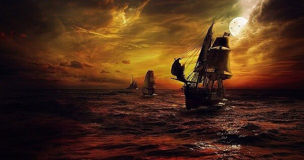 A dark orange and yellow tinted picture fading into black ocean scene with two ships sailing. The one in the foreground is a pirate ship and the smaller one is chasing after the pirate ship. In the background there is a lighthouse.