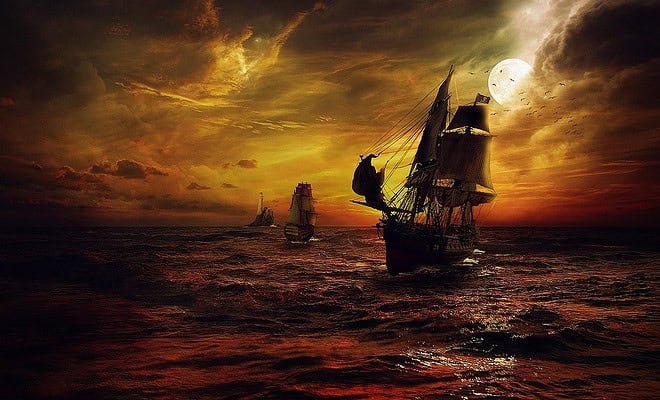 A dark orange and yellow tinted picture fading into black ocean scene with two ships sailing. The one in the foreground is a pirate ship and the smaller one is chasing after the pirate ship. In the background there is a lighthouse.