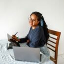 a lady sitting at a table and smiling in from of a laptop and tablet.
