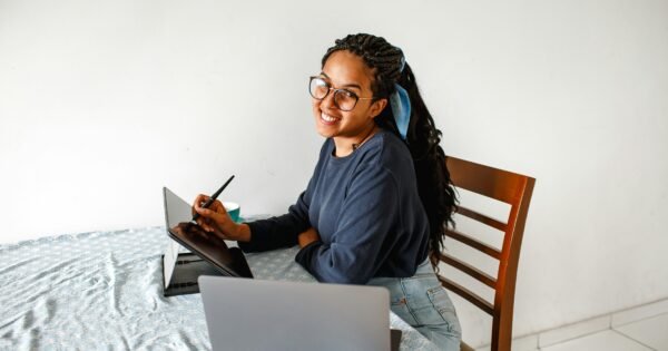 a lady sitting at a table and smiling in from of a laptop and tablet.
